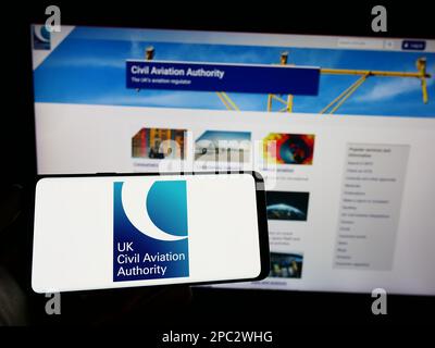 Person holding cellphone with logo of British regulator Civil Aviation Authority (CAA) on screen in front of webpage. Focus on phone display. Stock Photo