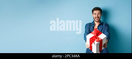Valentines day. Excited handsome man giving gift box and wishing happy holiday. Romantic guy extending hands with present, standing againt blue Stock Photo