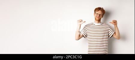 Confident young nerdy guy in glasses with red hair, pointing at himself and looking like professional, white background Stock Photo