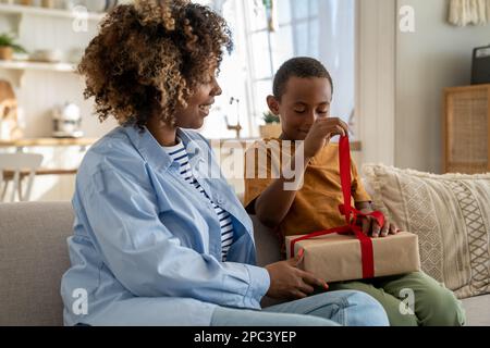 Pleased African American child boy happy to open gift from caring mom unties ribbon in anticipation Stock Photo