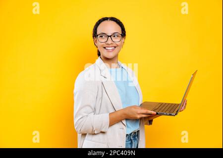 Happy successful stylish brazilian or hispanic business woman, company employee, consultant, with glasses, hold in hands open laptop, stand on isolated yellow background, looks aside, smiling Stock Photo