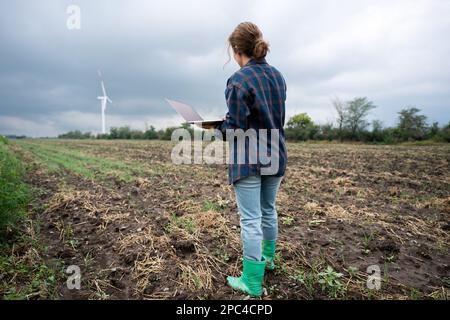 Farmer with laptop on the field. Wind turbines on a horizon. Smart sustainable farming and agriculture digitalization Stock Photo