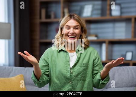 Close-up photo. Portrait of a young happy senior blonde woman sitting on the sofa at home wearing a green shirt. He smiles at the camera, enthusiastically spreads his hands, shows. Stock Photo