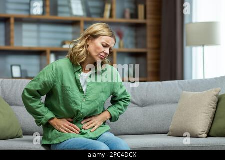 Unhealthy senior woman sitting on sofa at home. holding her stomach, grimacing in pain. She experiences severe abdominal pain, eating disorder, diarrhea, poisoning, menstruation, menopause. Stock Photo