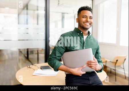 Carefree inspired indian male employee standing in modern office space and holding laptop, cheerful young businessman in green shirt looks aside with light friendly smile, full of plans and ideas Stock Photo