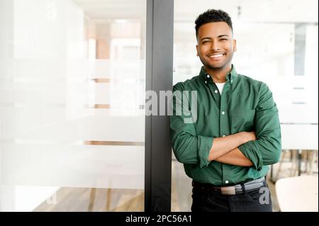 Successful indian authoritative confident young brunet adult male entrepreneur businessman professional in smart casual wear standing with arms crossed at the modern office, looking at the camera Stock Photo