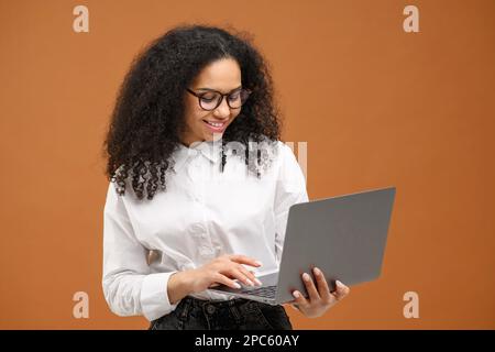 Purposeful African American woman entrepreneur or female office worker in formal shirt and glasses using laptop, white collar worker typing, websurfing, isolated on brown Stock Photo