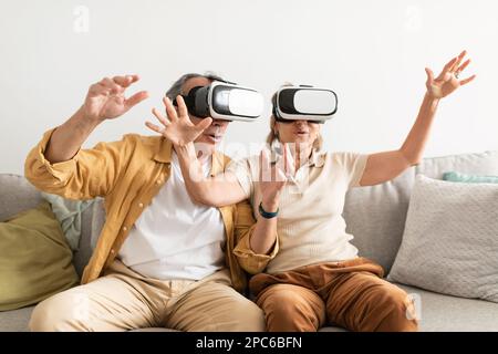 Senior couple having fun with virtual reality glasses, sitting on sofa and gesturing Stock Photo