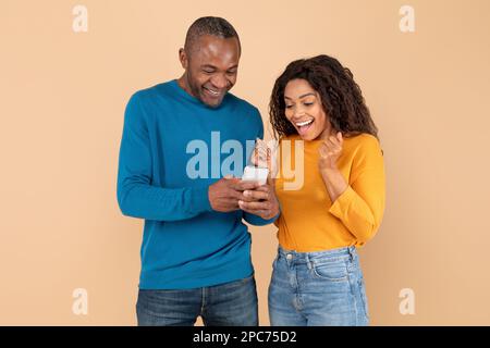 Wow. Excited black couple looking at cellphone screen, using new application on beige studio background Stock Photo