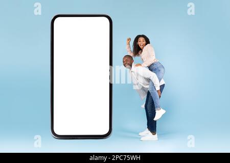 Great mobile app. Black couple having fun near giant smartphone with mockup copy space, man riding wife on back Stock Photo