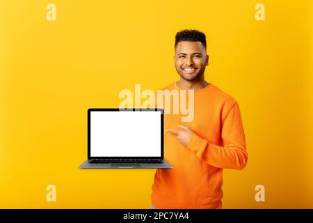 Positive optimistic indian guy freelancer or student showing laptop with empty screen and points finger on it, standing isolated on yellow, arab man presenting new app, mock-up, copy space Stock Photo
