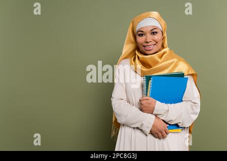 Cheerful multicultural student in hijab holding notebooks on green background,stock image Stock Photo