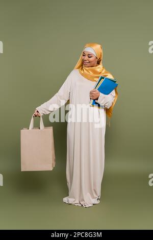 Carefree multiracial student in hijab holding shopping bags and notebooks on green background,stock image Stock Photo