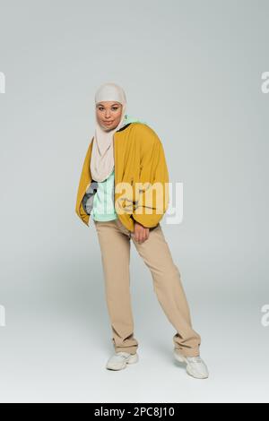 full length of multiracial woman in hijab and trendy casual outfit standing and smiling at camera on grey background,stock image Stock Photo