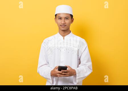 Young handsome asian muslim man smiling using smartphone standing over orange background Stock Photo