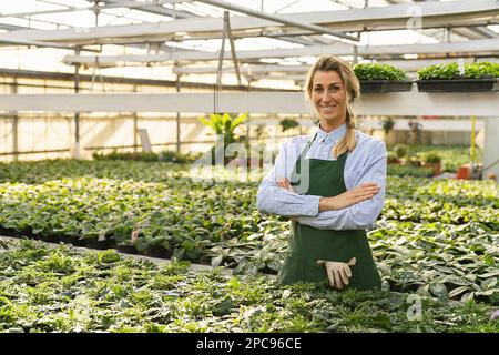 female florist posing in his garden center surrounded by green plants smiling to the camera joyfully with his arms crossed Stock Photo