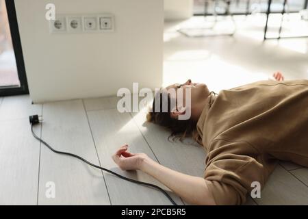 Woman lies on the floor with her eyes closed near electric cable with plug. Stock Photo