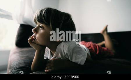 One small boy lying on couch with hand in chin hypnotized by entertainment media at home indoors. Lifestyle shot of kid watching cartoon Stock Photo