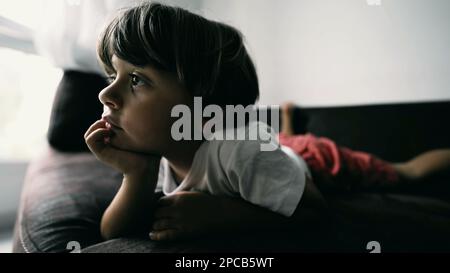 One small boy lying on couch with hand in chin hypnotized by entertainment media at home indoors. Lifestyle shot of kid watching cartoon Stock Photo
