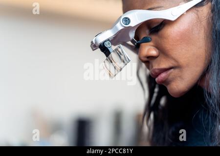 Side view of crop ethnic woman wearing headband with magnifying glass while working in modern beauty salon as microblading master Stock Photo