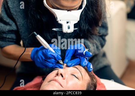 Professional microblading master in latex gloves and magnification goggles applying eyebrow tattoo to female client Stock Photo