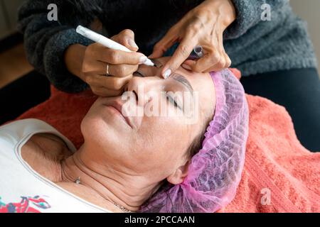 From above of crop anonymous female cosmetologist preparing mature woman for eyebrow permanent makeup procedure in beauty salon Stock Photo