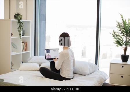 Back view of darked haired woman sitting on white bed and browsing website with trendy clothes on sale. Caucasian female using wireless laptop for cho Stock Photo