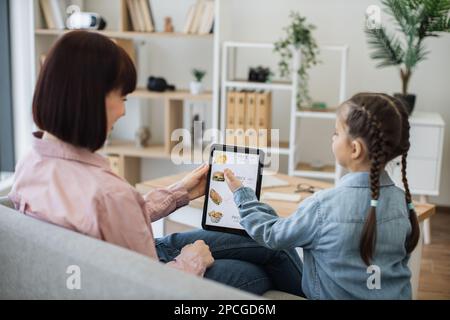 Brunette caucasian ladies surfing fast food delivery online website using digital tablet while staying in apartment. Beautiful woman and cute girl checking available discounts on snacks indoors. Stock Photo