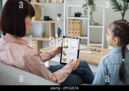 Brunette caucasian ladies surfing fast food delivery online website using digital tablet while staying in apartment. Beautiful woman and cute girl checking available discounts on snacks indoors. Stock Photo