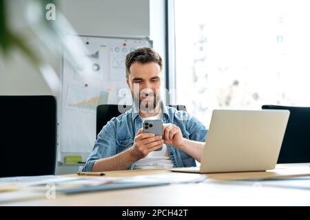 Positive caucasian man, freelancer, programmer, seo, sits at a table in the office, uses a smartphone, messaging on social networks, conducts work correspondence, watches news, smiles Stock Photo