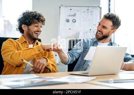 Success, deal. Cheerful colleagues, indian and caucasian men, sitting in a creative office, working on a new project, a startup, developing a strategy and deadlines, happy with the result, smiling Stock Photo