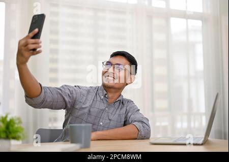 Handsome and smiling millennial Asian man or businessman taking selfie with his phone while sitting at his office desk. Stock Photo