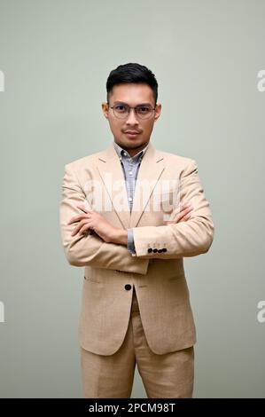 Portrait of determined and professional adult Asian businessman in formal business suit stands with arms crossed against green background. Stock Photo