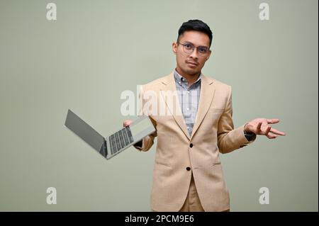 Confused and doubtful millennial Asian businessman in formal business suit stands against a green studio background with his laptop. Stock Photo