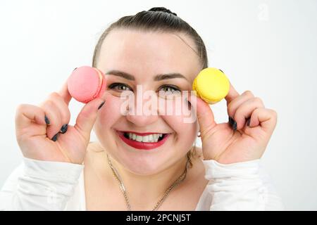 a fat insatiable woman smiles happily holding two macaroons of different colors in hands put dessert on her face, the food is sweet and harmful Stock Photo
