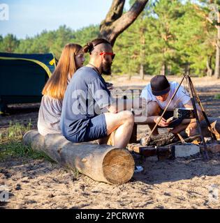 A company of friends of girls and guys kindle a fire against the backdrop of the beautiful nature of the forest and the river bank. Hiking, outdoor re Stock Photo