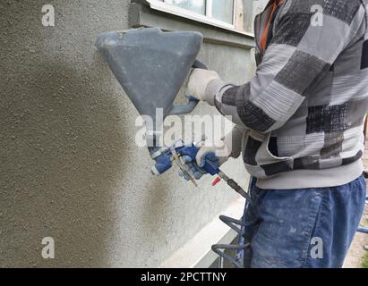 A builder is rendering, plastering, coating house  exterior wall of a building using plaster sprayer machine, stucco concrete sprayer gun. Stock Photo