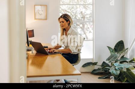 Female entrepreneur typing on a laptop keyboard while sitting in her office. Focused young businesswoman writing an email to her business clients. Stock Photo