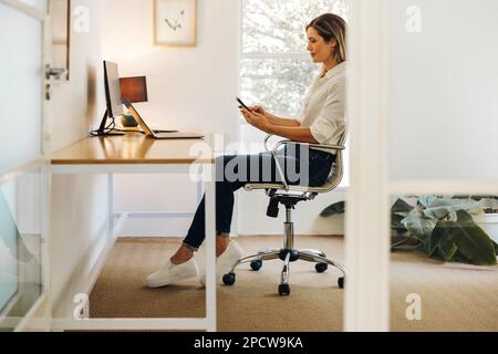 Cheerful businesswoman using a smartphone while working in her office. Young female entrepreneur communicating with her business clients online. Stock Photo