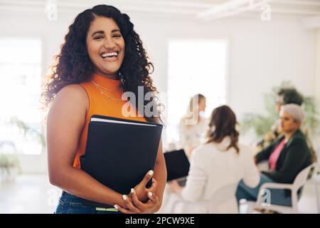 Happy young businesswoman smiling at the camera while standing in a boardroom. Cheerful young businesswoman attending a meeting with her team in an al Stock Photo