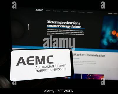 Person holding cellphone with logo of Australian Energy Market Commission (AEMC) on screen in front of webpage. Focus on phone display. Stock Photo