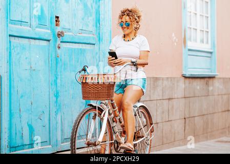 One young mature woman in outdoor leisure activity riding a bike and using mobile phone outside home in the street. Spring active green transport bicy Stock Photo