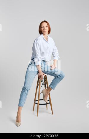 Casual portrait of middle aged redhead woman in white shirt and blue jeans sitting on tall stool and stretching one leg to the side Stock Photo