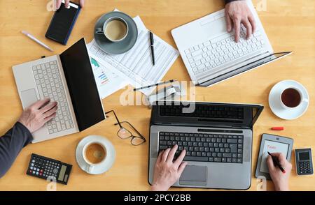 Working together on a joint business project, top view of the wooden table with hands, three laptops, papers, calculator and coffee, high angle shot f Stock Photo