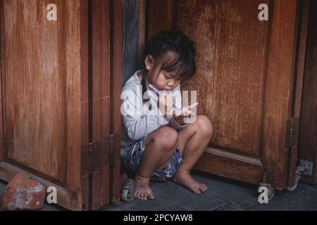 Pattaya, Thailand - December 21, 2022: A sweet Asian little girl,  little asian girl playing with herself in front of wooden door, Pattaya Floating Ma Stock Photo