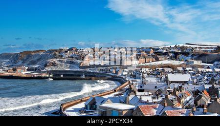 Cullen Bay Moray Scotland blue sky snow covered houses and waves breaking on the beach and barrier walls Stock Photo