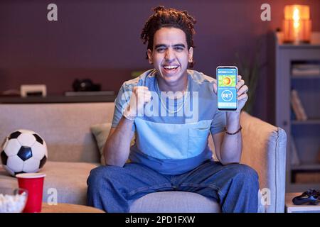 Portrait of excited young man watching sports match on TV at home and using online betting app in blue light, copy space Stock Photo
