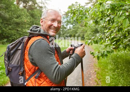 Portrait of happy elderly man with hiking poles and backpack standing in forest during vacation Stock Photo