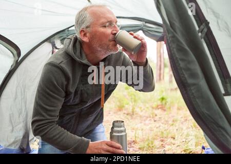 Senior man in tent drinking cup of hot coffee from thermos jug in nature Stock Photo