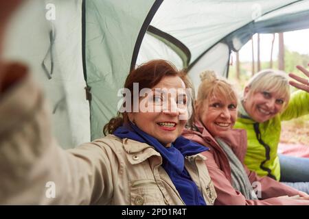 Happy elderly woman taking selfie with female friends sitting in camping tent at forest Stock Photo